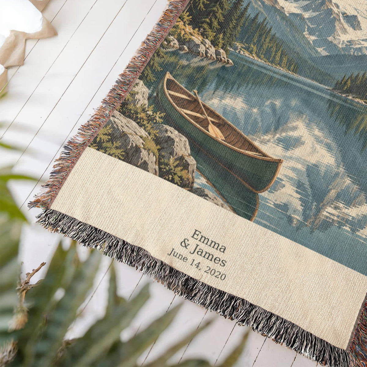 Mountain Lake woven cotton blanket laid at an angle on a white wood floor beside a plant, showing the canoe, the knotted fringe and "Emma & James, June 14, 2020" on the cream panel.
