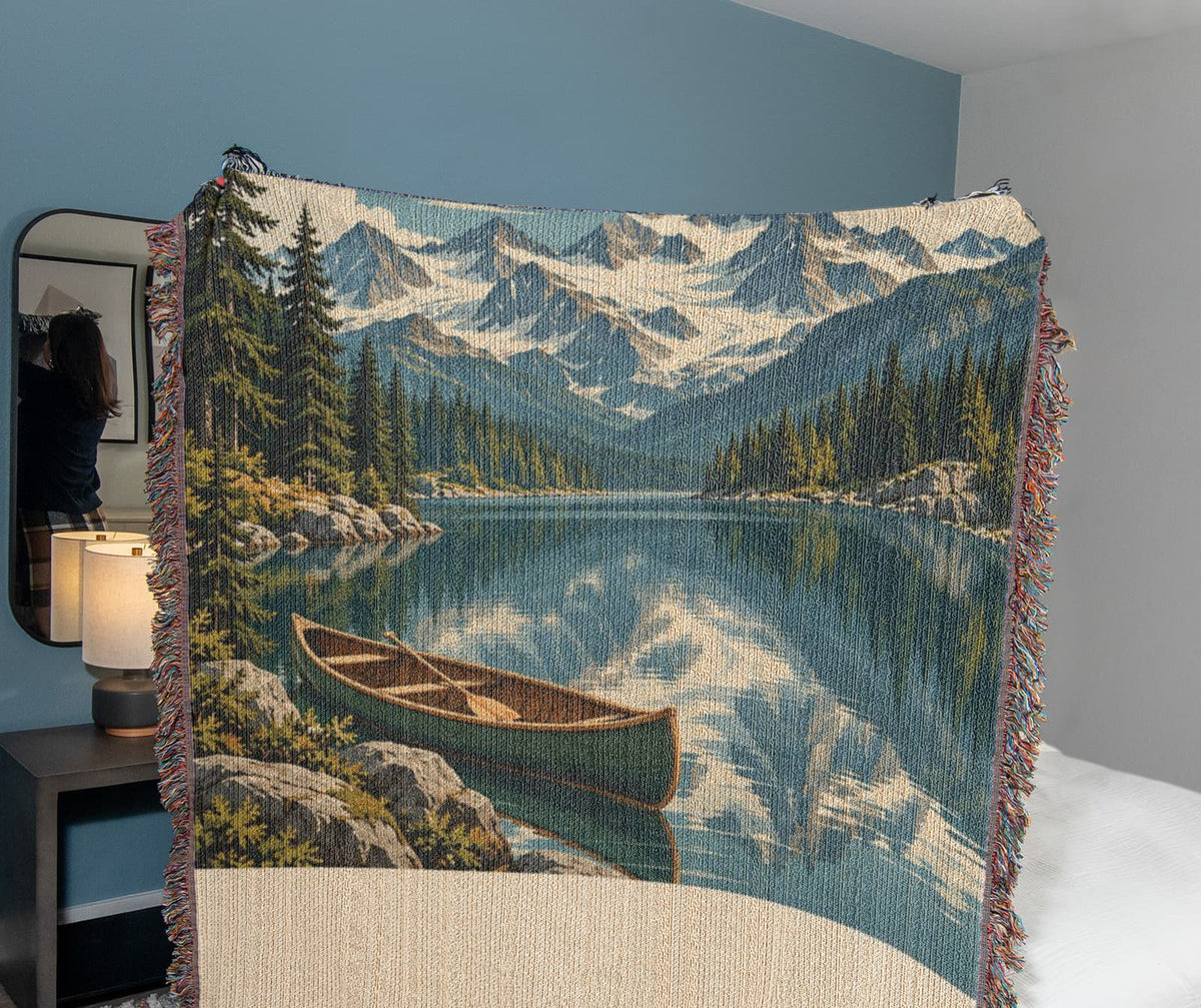 Mountain Lake woven cotton blanket draped in a cosy bedroom, the woven mountains, lake reflection and canoe artwork in full view.