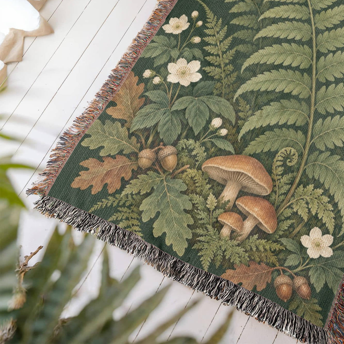 Close-up of a corner of the Woodland Garden woven cotton blanket lying at an angle on a white wood floor, showing the knotted fringe, woven texture and ferns and woodland plants.