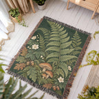 Woodland Garden woven blanket laid flat on a white wooden floor in a sunny room, showing tall green ferns, white flowers, mushrooms and autumn leaves on deep green framed by fringe.