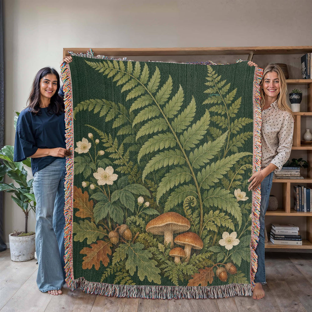 Two women holding up the Woodland Garden woven cotton blanket in a bright living room, showing tall green ferns, white flowers, mushrooms and autumn leaves on deep green and the woven texture.