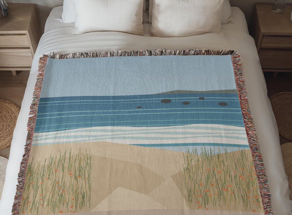 Coastal woven cotton blanket laid across a made bed between two nightstands, the woven sea, surf and dune grass artwork in full view.