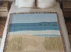 Coastal woven cotton blanket laid across a made bed between two nightstands, the woven sea, surf and dune grass artwork in full view.