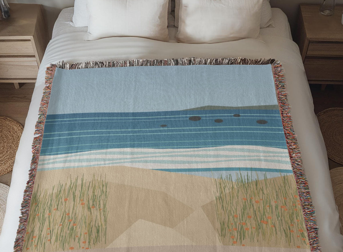 Coastal woven cotton blanket laid across a made bed between two nightstands, the woven sea, surf and dune grass artwork in full view.