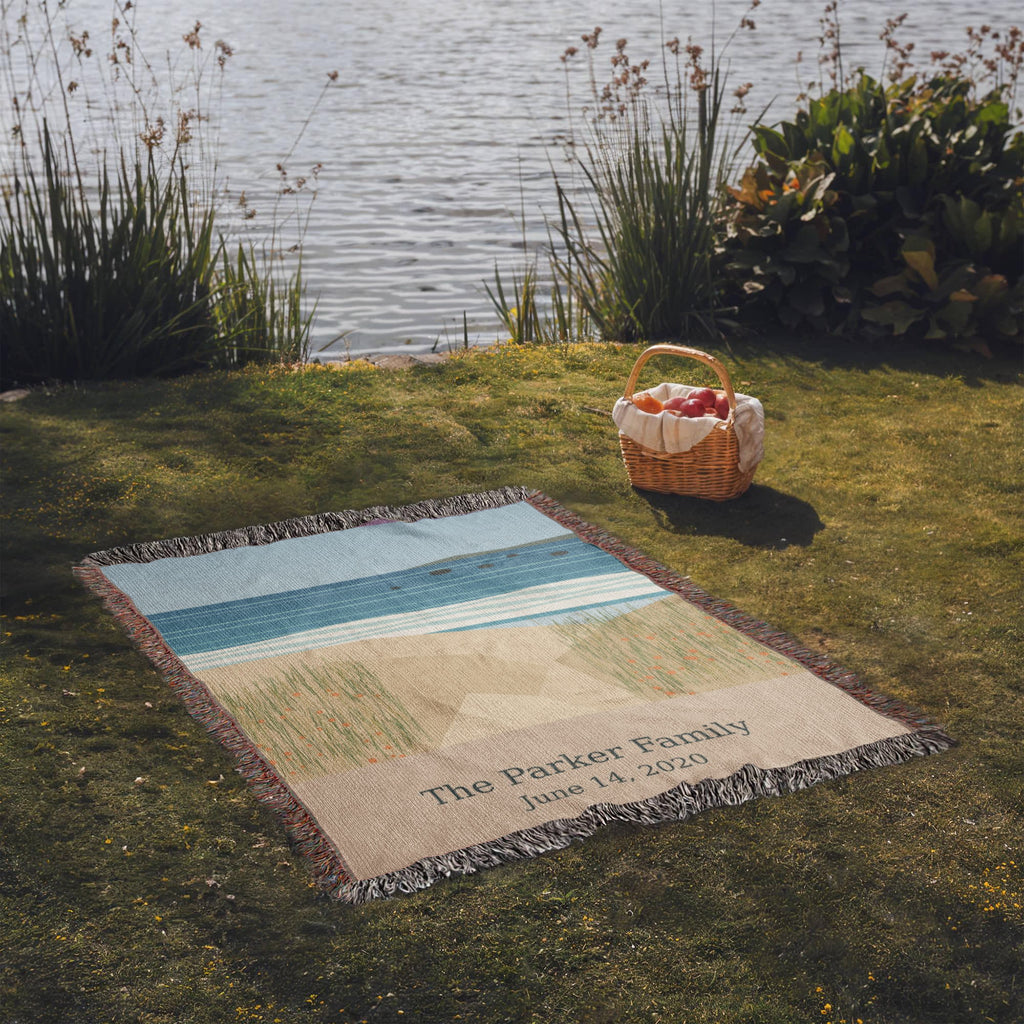 Coastal personalized woven cotton blanket spread on lakeside grass beside a picnic basket, the whole blanket in view.