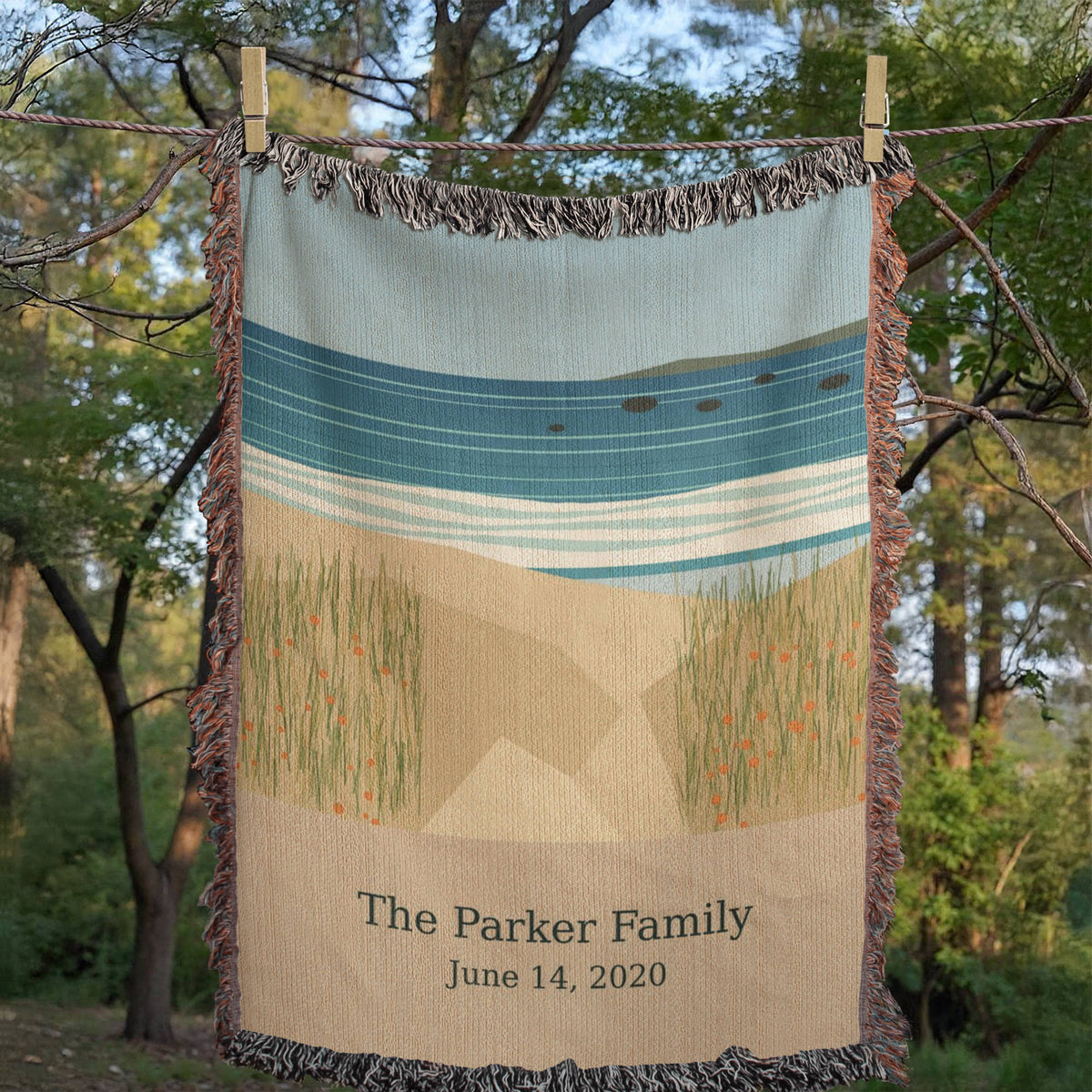 Coastal personalized woven cotton blanket pegged on a clothesline among trees, showing "The Parker Family" and June 14, 2020 on the sand panel.