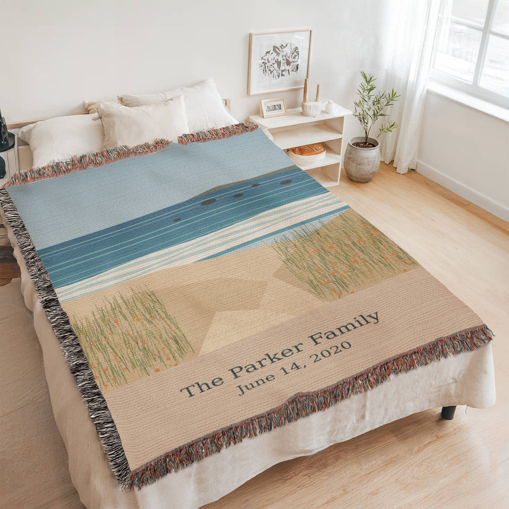 Coastal personalized woven cotton blanket spread across a bed in a bright bedroom, seen at an angle with the whole blanket in frame.