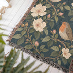 Close-up of a corner of the Bird Garden woven cotton blanket lying at an angle on a white wood floor, showing the knotted fringe, woven texture and wild roses and branches.