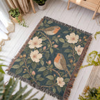Bird Garden woven blanket laid flat on a white wooden floor in a sunny room, showing robins perched on flowering branches with cream wild roses on deep teal framed by fringe.