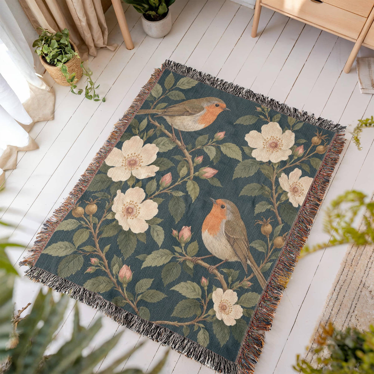 Bird Garden woven blanket laid flat on a white wooden floor in a sunny room, showing robins perched on flowering branches with cream wild roses on deep teal framed by fringe.