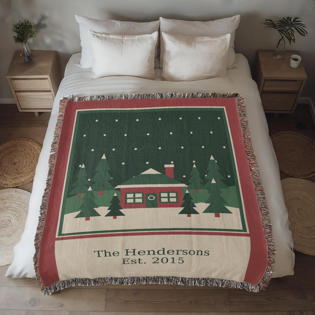 Personalized family name Christmas woven cotton blanket spread on a bed, showing the red cabin, evergreens, family name and year with fringe