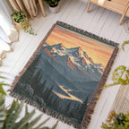 Mountain Sunrise woven blanket laid flat on a white wooden floor beside a plant and a wooden bench, the whole design and knotted fringe in view.