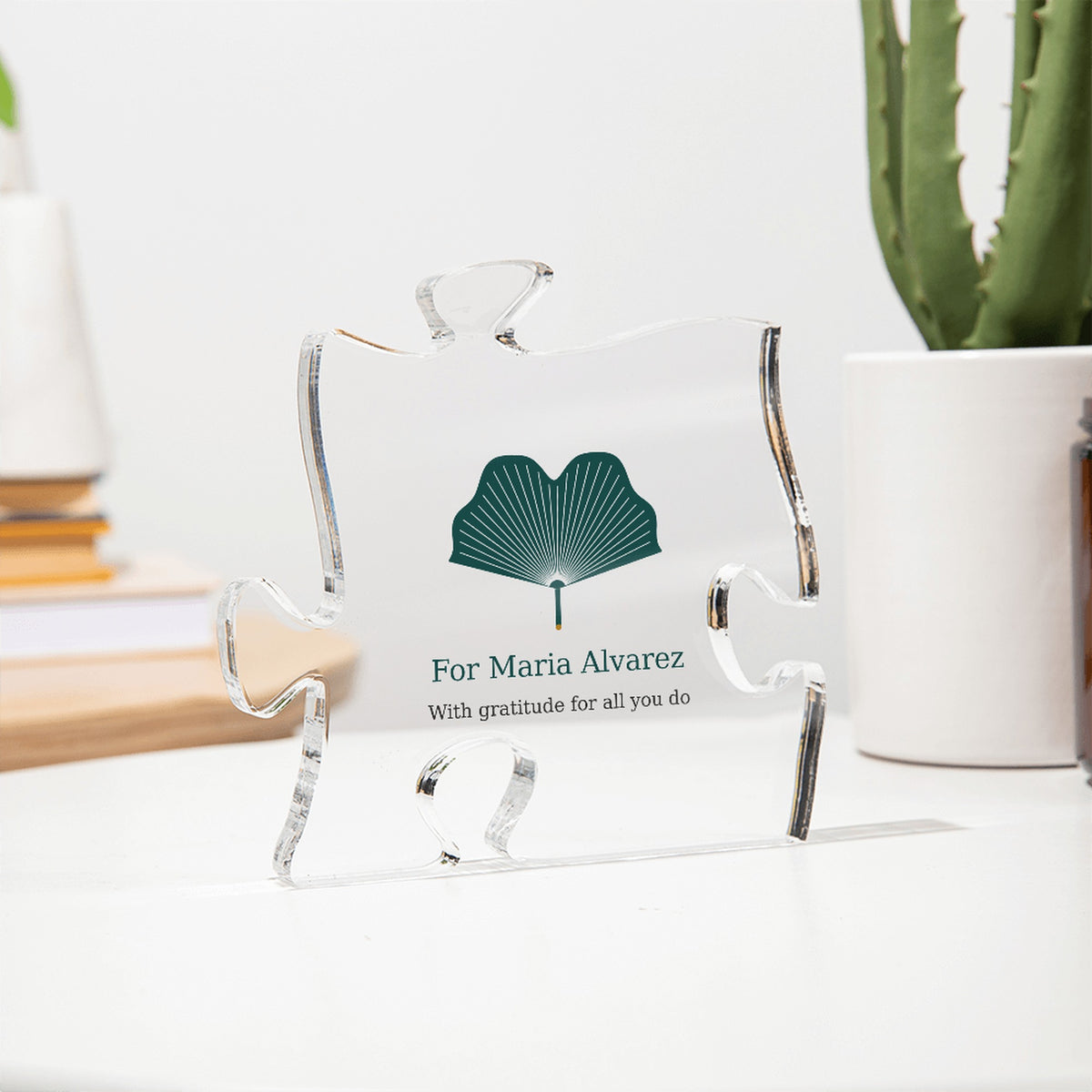 Clear acrylic puzzle-piece caregiver plaque seen at an angle on a white desk beside a cactus, with the text "For Maria Alvarez" and "With gratitude for all you do"