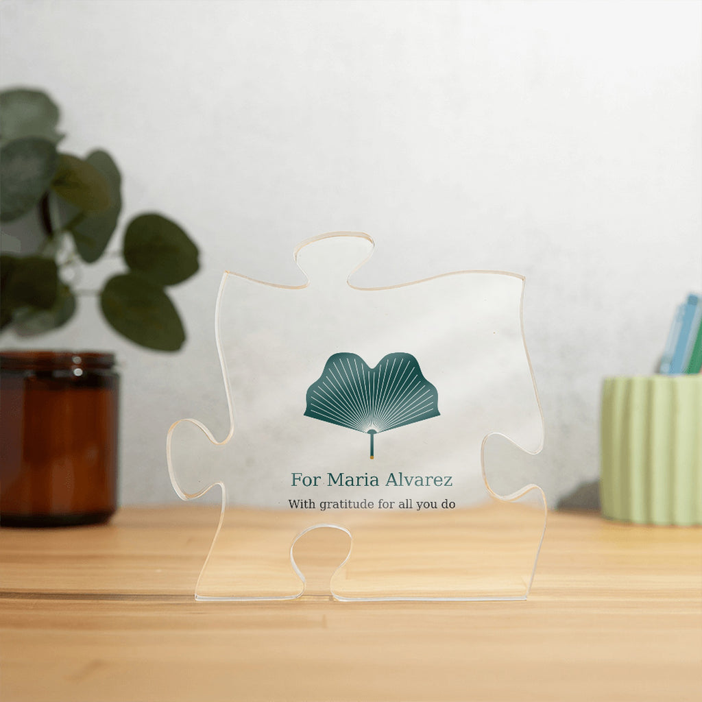 Clear acrylic puzzle-piece caregiver plaque on a wooden surface with plants, with the text "For Maria Alvarez" and "With gratitude for all you do"