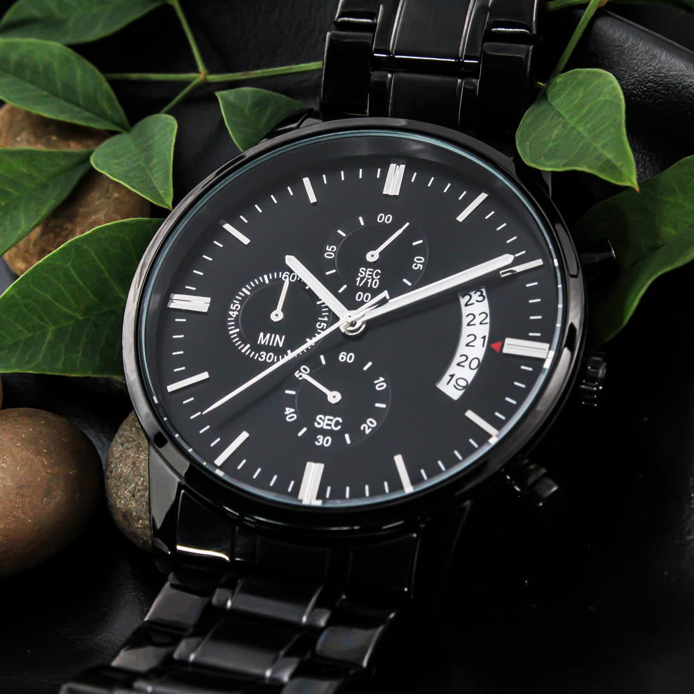 Black chronograph watch dial with three sub-dials and a date window, among green leaves and stones