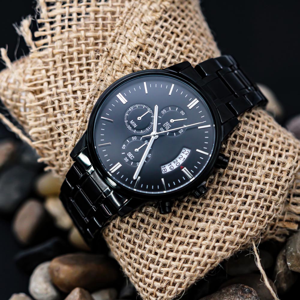 Black chronograph watch with a black metal bracelet resting on burlap with stones
