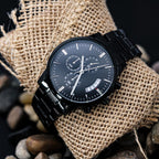 Black chronograph watch with a black metal bracelet resting on burlap with stones