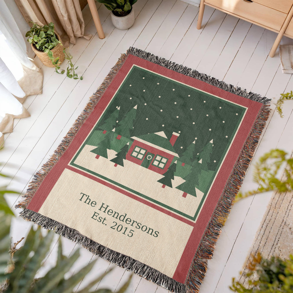 Christmas woven cotton blanket laid at an angle on a white wood floor, showing the woven cabin scene, family name and year