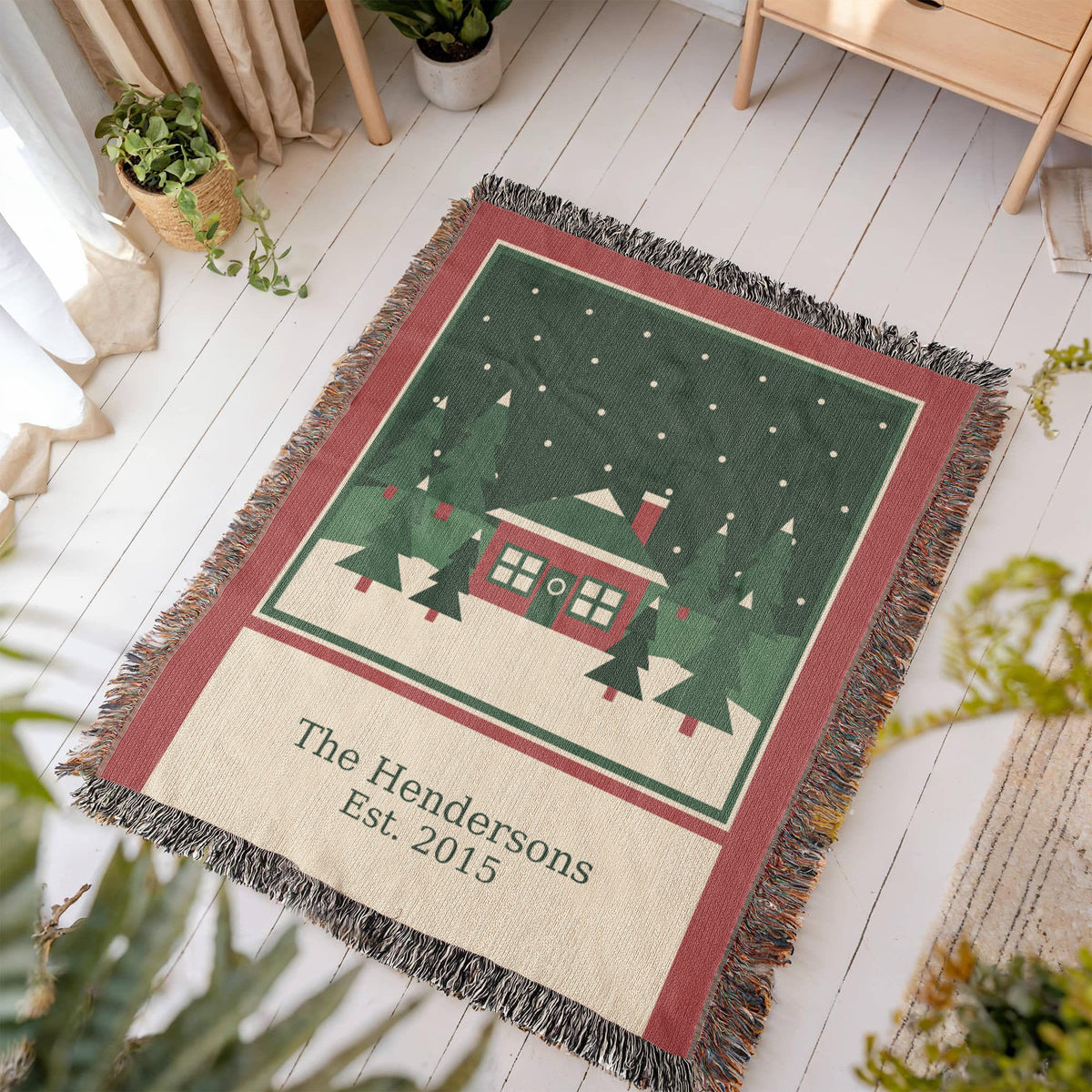 Christmas woven cotton blanket laid at an angle on a white wood floor, showing the woven cabin scene, family name and year