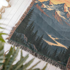 Close-up of a corner of the Mountain Sunrise woven cotton blanket lying at an angle on a white wood floor, showing the knotted fringe, woven texture and pine forest and valley.