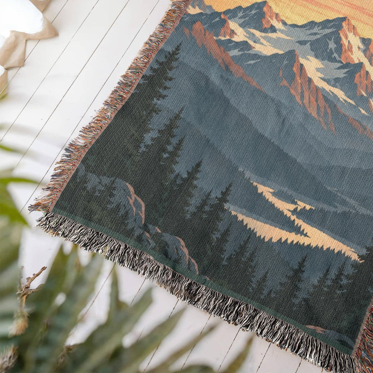 Close-up of a corner of the Mountain Sunrise woven cotton blanket lying at an angle on a white wood floor, showing the knotted fringe, woven texture and pine forest and valley.