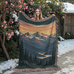 Mountain Sunrise woven blanket held up by a woman in a flowering garden, showing the orange sky, snowy peaks and pine valley with fringed edges.