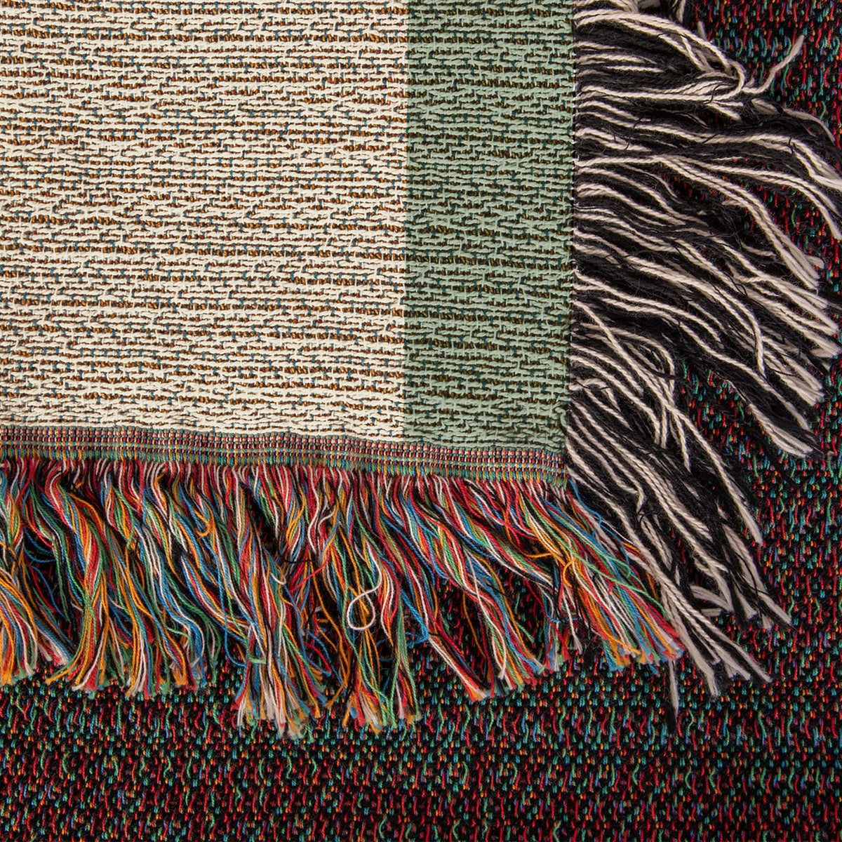 Close-up of the woven cotton blanket corner: textured cream weave, sage border and multicolor knotted fringe