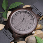 Front view of a dark wooden watch with a black leather strap among green leaves and stones