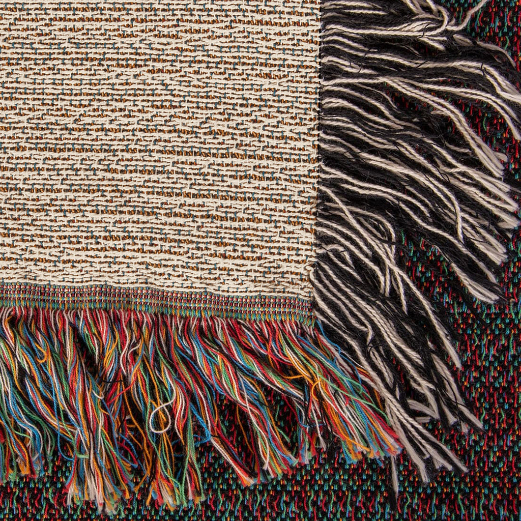 Corner close-up of the Family Caravan woven cotton blanket showing the cream cotton weave, the multicolour bottom fringe and the black-and-white side fringe.