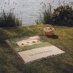 Family Caravan personalized woven cotton blanket spread on lakeside grass beside a picnic basket, the whole blanket in view.