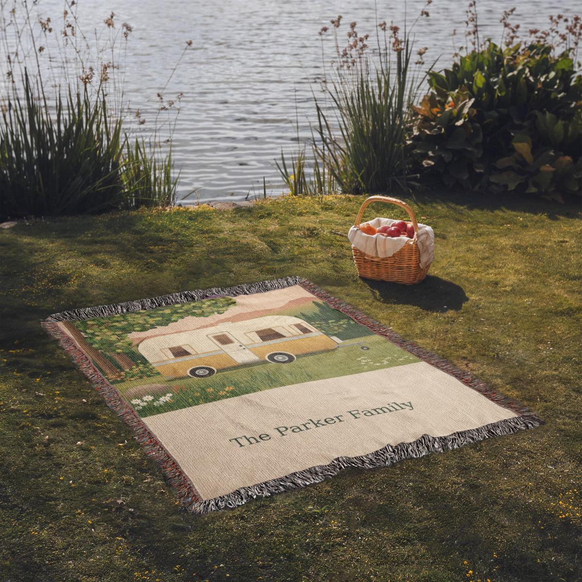 Family Caravan personalized woven cotton blanket spread on lakeside grass beside a picnic basket, the whole blanket in view.
