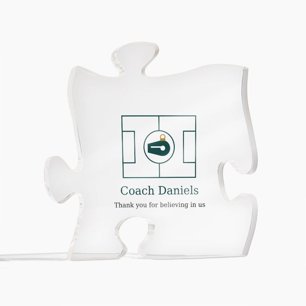 Clear acrylic puzzle-piece coach plaque on a white background, printed with a green design and the text "Coach Daniels" and "Thank you for believing in us"
