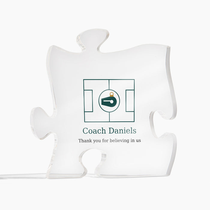 Clear acrylic puzzle-piece coach plaque on a white background, printed with a green design and the text "Coach Daniels" and "Thank you for believing in us"