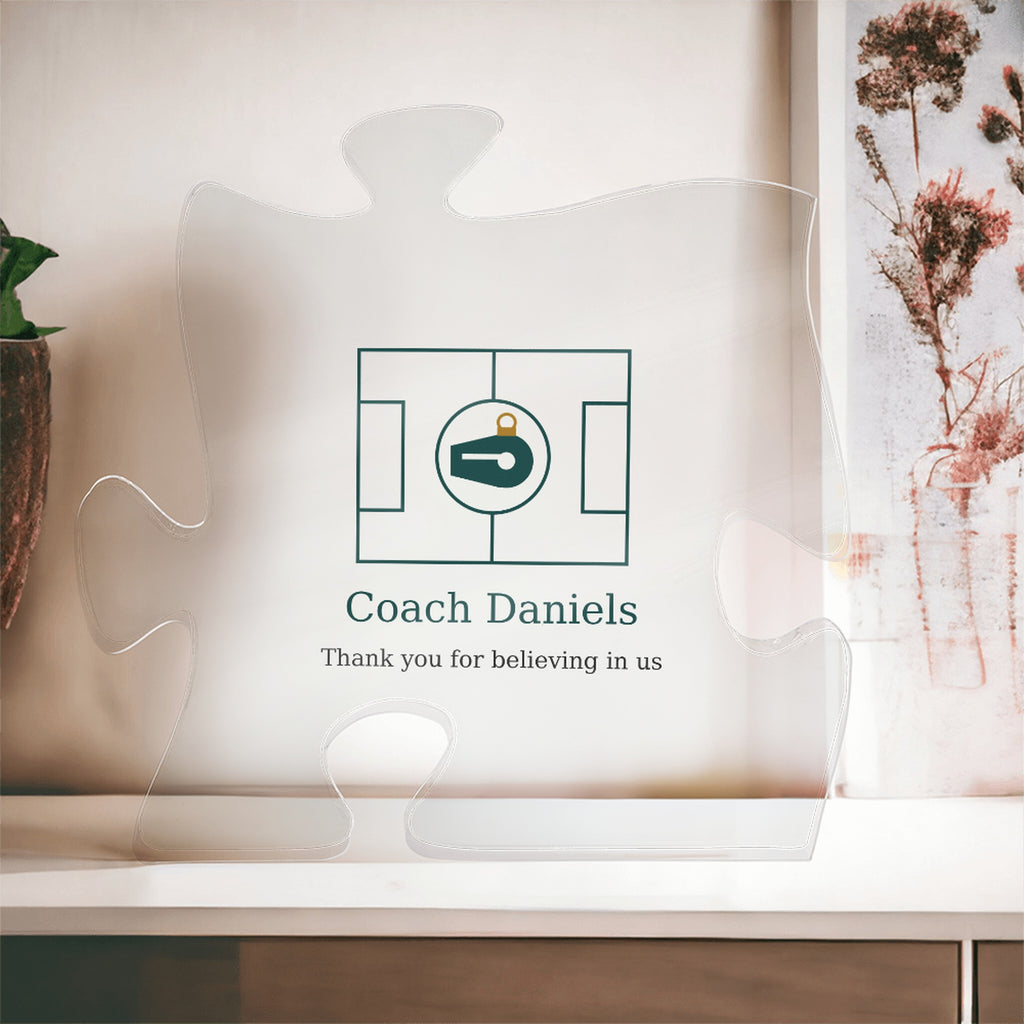 Clear acrylic puzzle-piece coach plaque in front of a window with dried flowers, with the text "Coach Daniels" and "Thank you for believing in us"