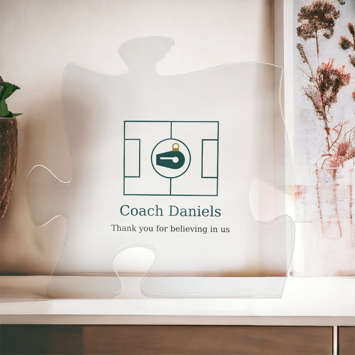 Clear acrylic puzzle-piece coach plaque in front of a window with dried flowers, with the text "Coach Daniels" and "Thank you for believing in us"