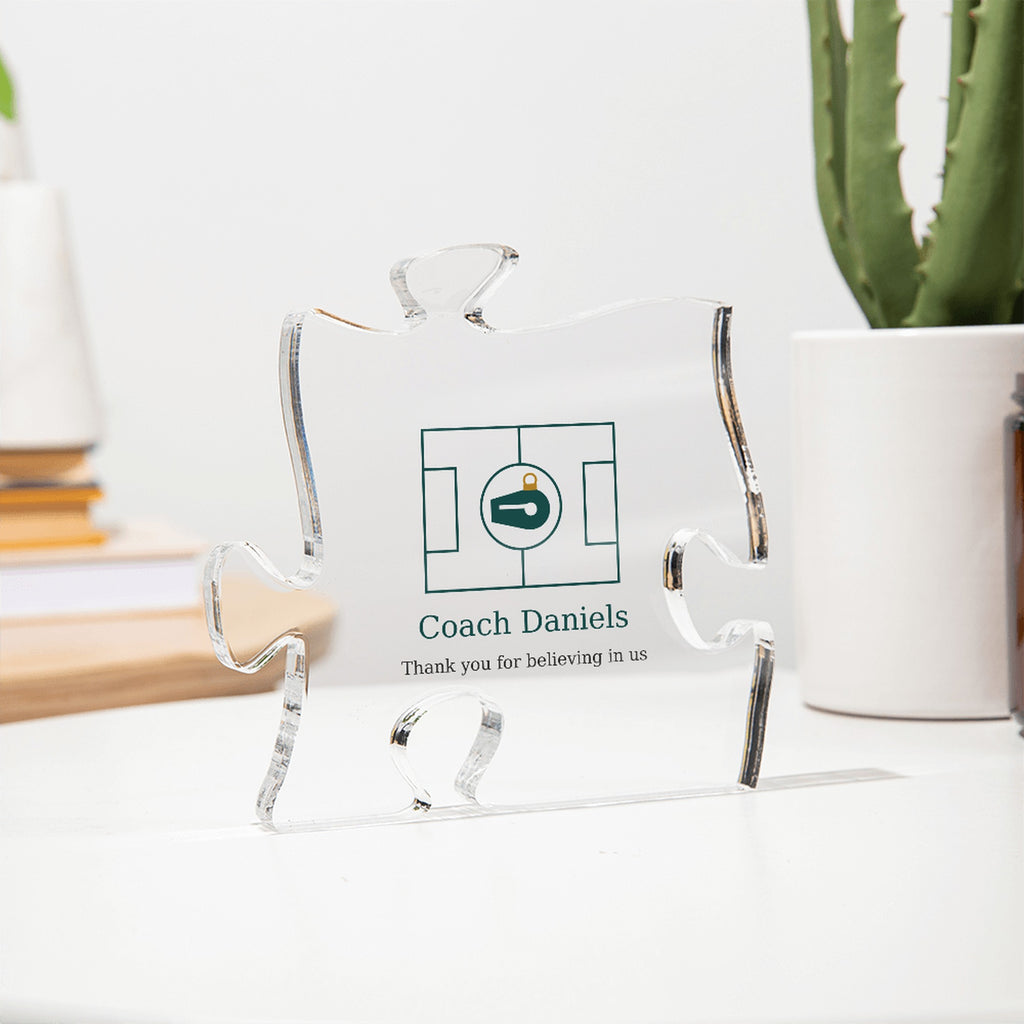 Clear acrylic puzzle-piece coach plaque seen at an angle on a white desk beside a cactus, with the text "Coach Daniels" and "Thank you for believing in us"