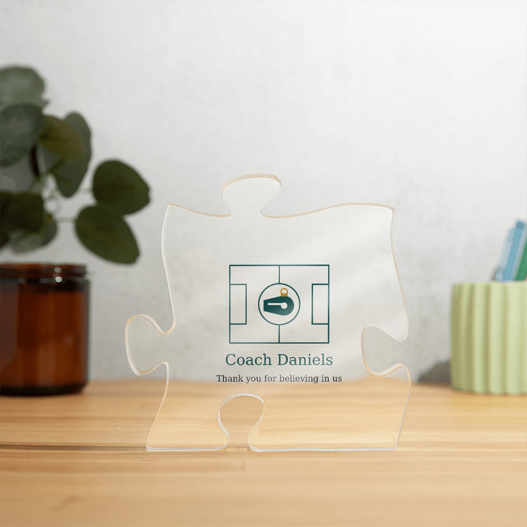 Clear acrylic puzzle-piece coach plaque on a wooden surface with plants, with the text "Coach Daniels" and "Thank you for believing in us"