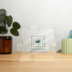 Clear acrylic puzzle-piece coach plaque on a wooden surface with plants, with the text "Coach Daniels" and "Thank you for believing in us"
