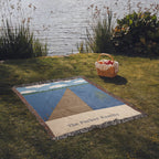 Lake House personalized woven cotton blanket spread on lakeside grass beside a picnic basket, the whole blanket in view.