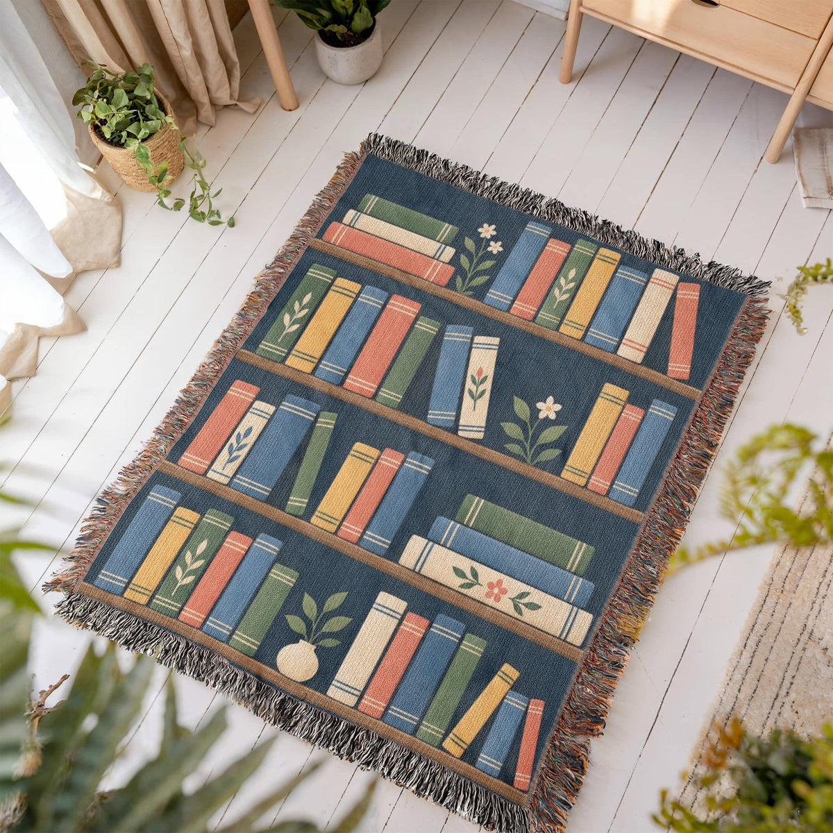 Book Garden woven blanket laid flat on a white wooden floor in a sunny room, showing shelves of colorful books with small potted plants on navy framed by fringe.