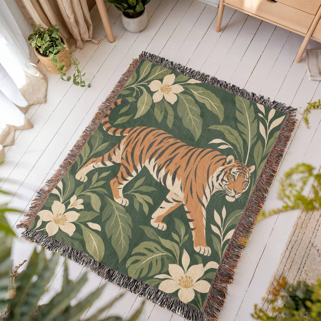Tiger Botanical woven blanket laid flat on a white wooden floor in a sunny room, showing a striding orange tiger among cream blossoms and green leaves on deep green framed by fringe.