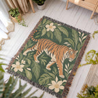 Tiger Botanical woven blanket laid flat on a white wooden floor in a sunny room, showing a striding orange tiger among cream blossoms and green leaves on deep green framed by fringe.