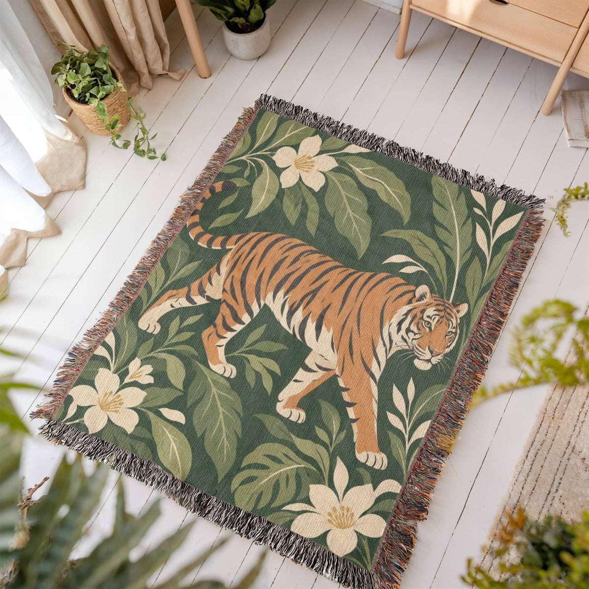 Tiger Botanical woven blanket laid flat on a white wooden floor in a sunny room, showing a striding orange tiger among cream blossoms and green leaves on deep green framed by fringe.
