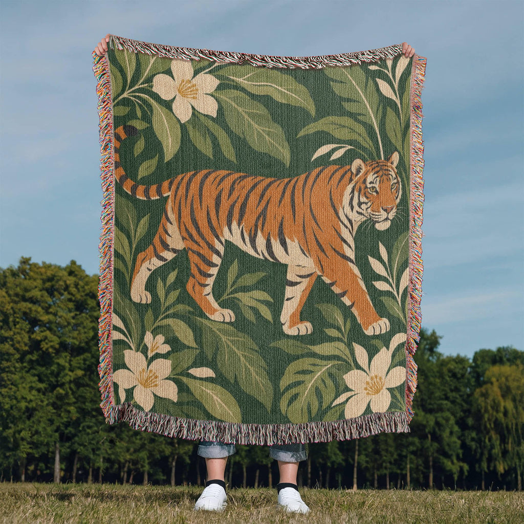 Tiger Botanical woven blanket held up outdoors against a blue sky over a grassy field, showing a striding orange tiger among cream blossoms and green leaves on deep green with fringed edges.