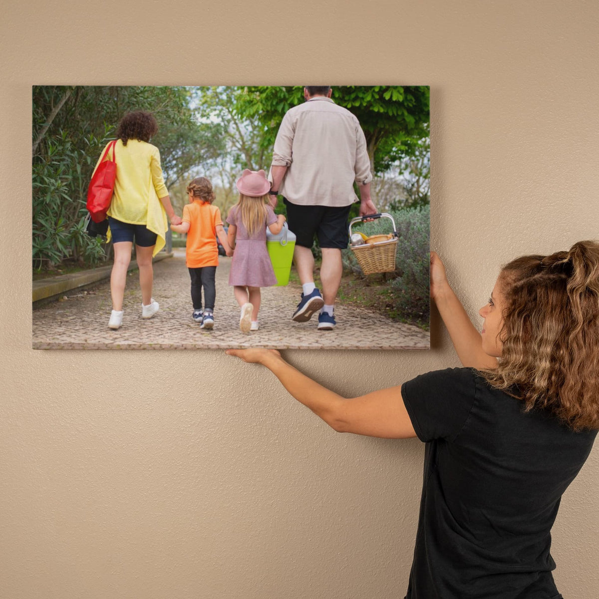 Personalized family photo canvas being hung on a warm beige wall, showing parents and two children carrying a picnic basket down a leafy path.