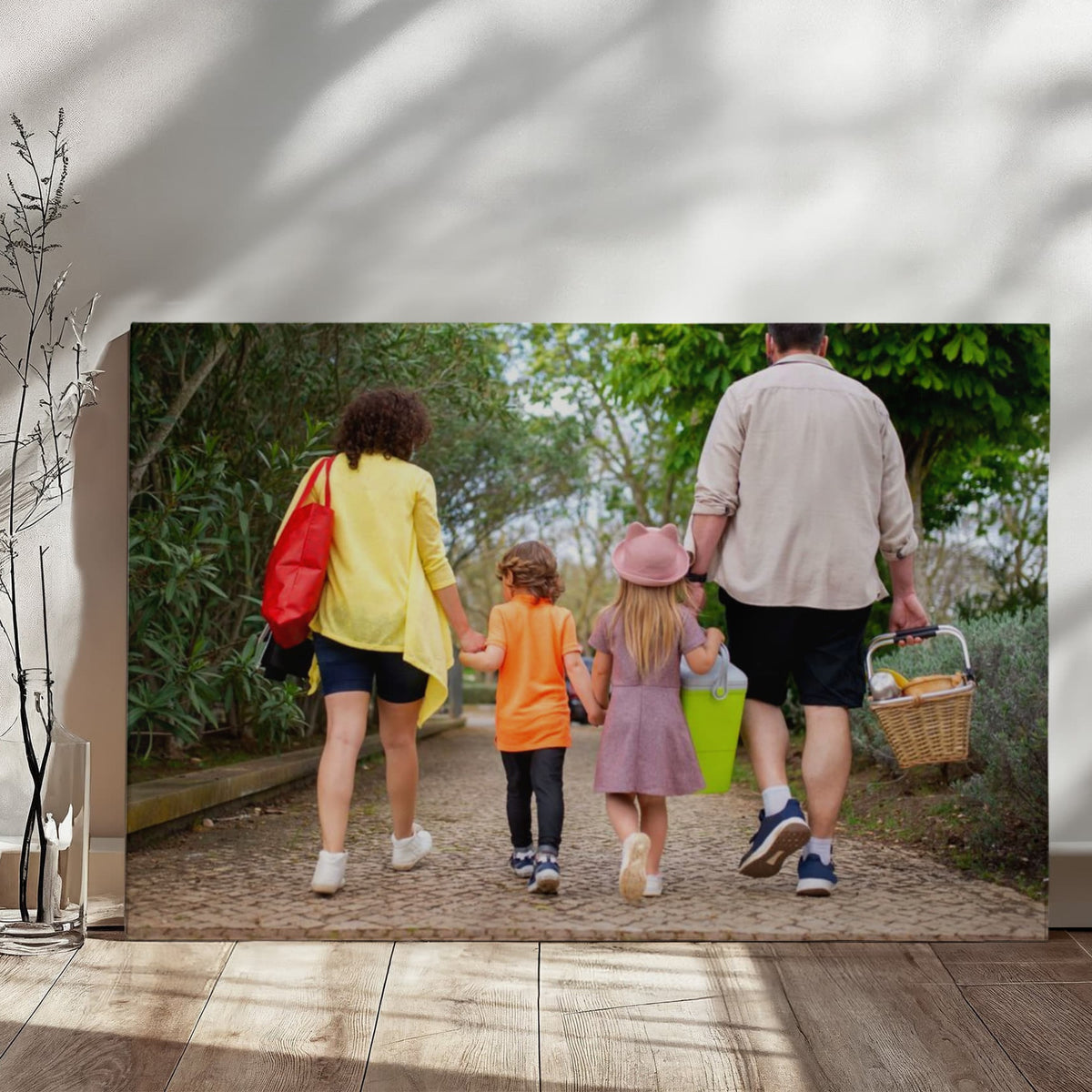 Gallery-wrapped family canvas leaning against a sunlit white wall on a wooden floor, with soft window shadows and a dried branch beside it.
