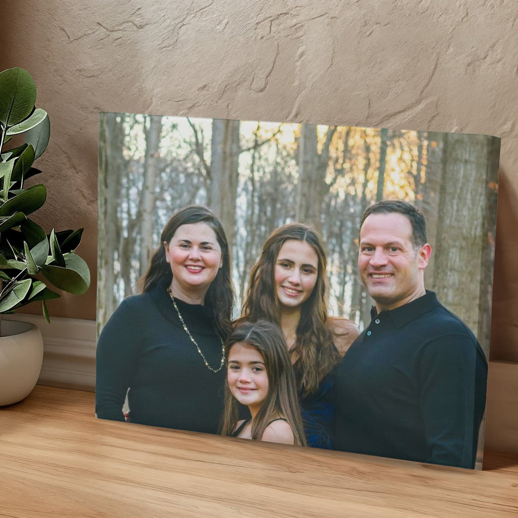 Personalized family photo canvas leaning against a textured plaster wall on a wooden floor, next to a leafy green plant in a white pot.