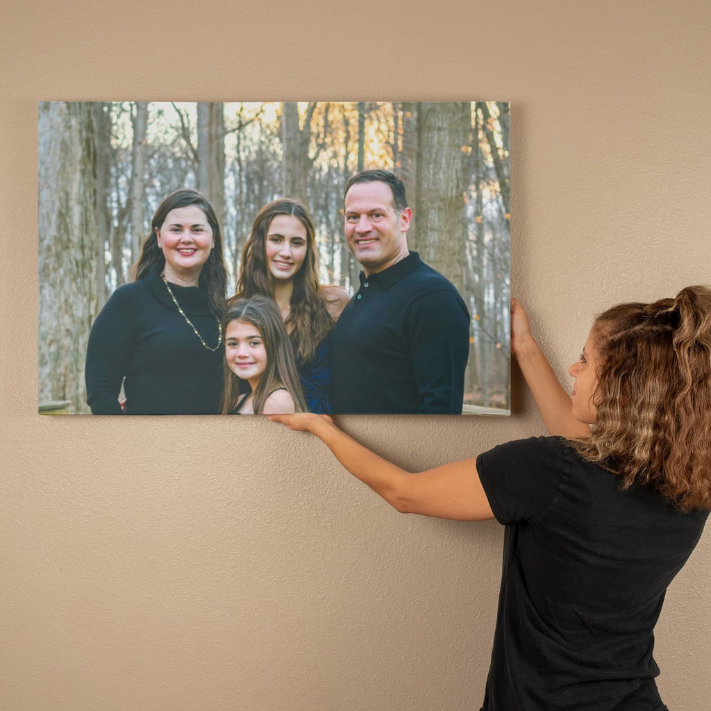 Woman hanging a custom family portrait canvas on a warm beige wall, showing parents and two daughters smiling among autumn trees.